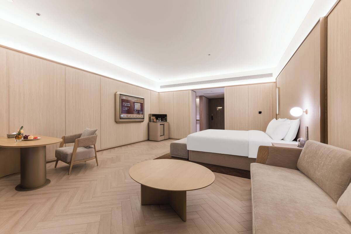 A modern hotel room with a bed, a sofa, a round coffee table, a desk, and a chair.