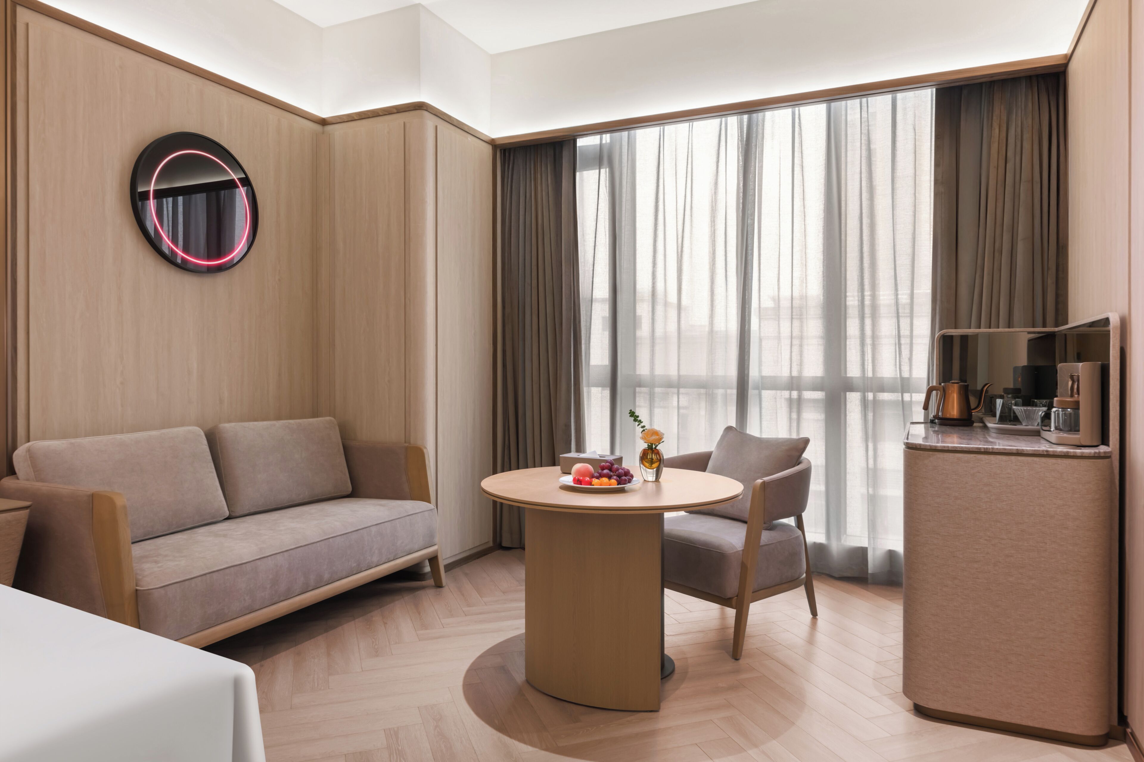 Una habitación de hotel moderna con una mesa redonda, un sofá, una silla, un minibar y un ventanal con cortinas.