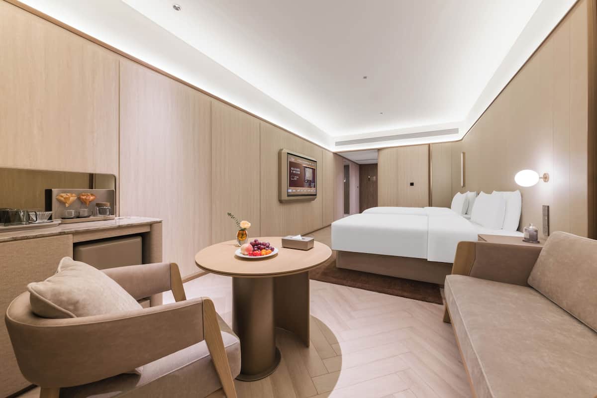 A modern hotel room with a bed, a round table with a fruit bowl, a sofa, and a desk with a computer.
