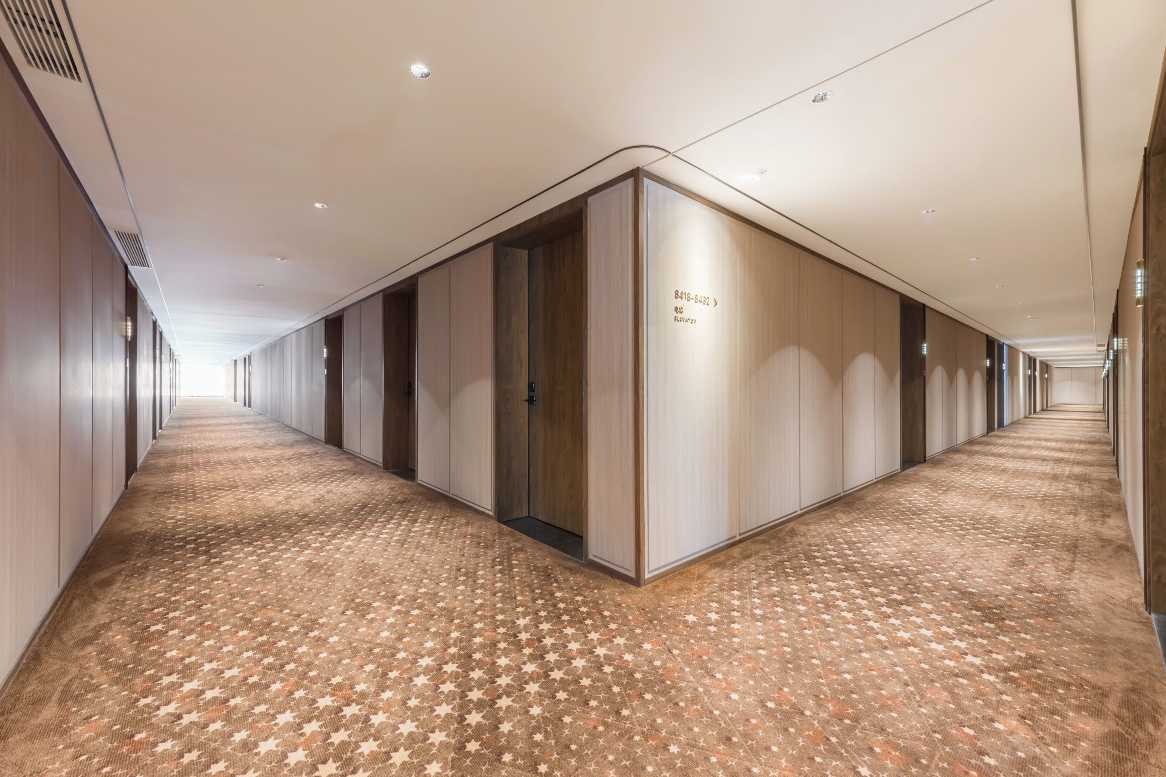 A long, narrow hotel corridor with a patterned carpet, wooden walls, and recessed lighting.