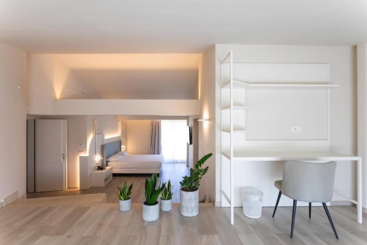 A modern, well-lit room with a bed, bedside table, and a desk with a chair. There are potted plants on the floor.