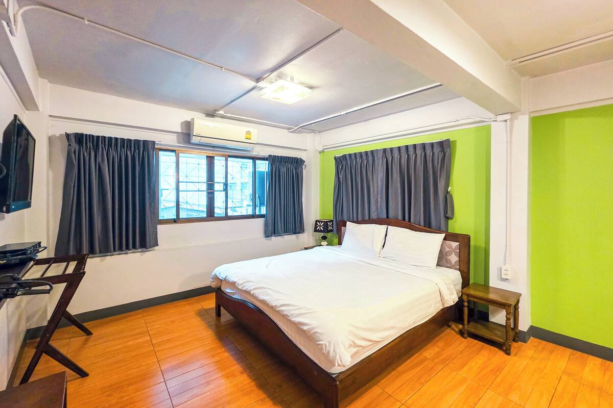 A hotel room with a bed, a desk, a chair, a window with curtains, and a green wall.
