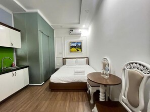 A compact hotel room with a bed, a small table, a chair, a kitchenette, and a picture on the wall. - StayX Saigon Centre (Xuan Hoa)