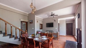 Un comedor con una mesa y sillas de madera, una escalera, un ventilador de techo y un televisor colgado en la pared.