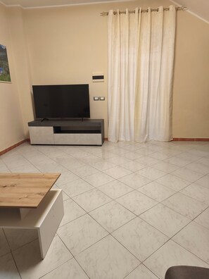 A living room with a flat-screen TV on a stand, a wooden table, and a curtain. - Cibali rooms (Catania)