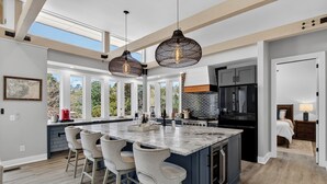 A modern kitchen with a large island, bar stools, pendant lighting, and a view through large windows. - Private Pool + Outdoor Lounge + Movie Room + Game Room | Summerset by AvantStay (Isle Of Palms)