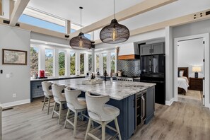 A modern kitchen with a large island, bar stools, pendant lighting, and a view through large windows.
