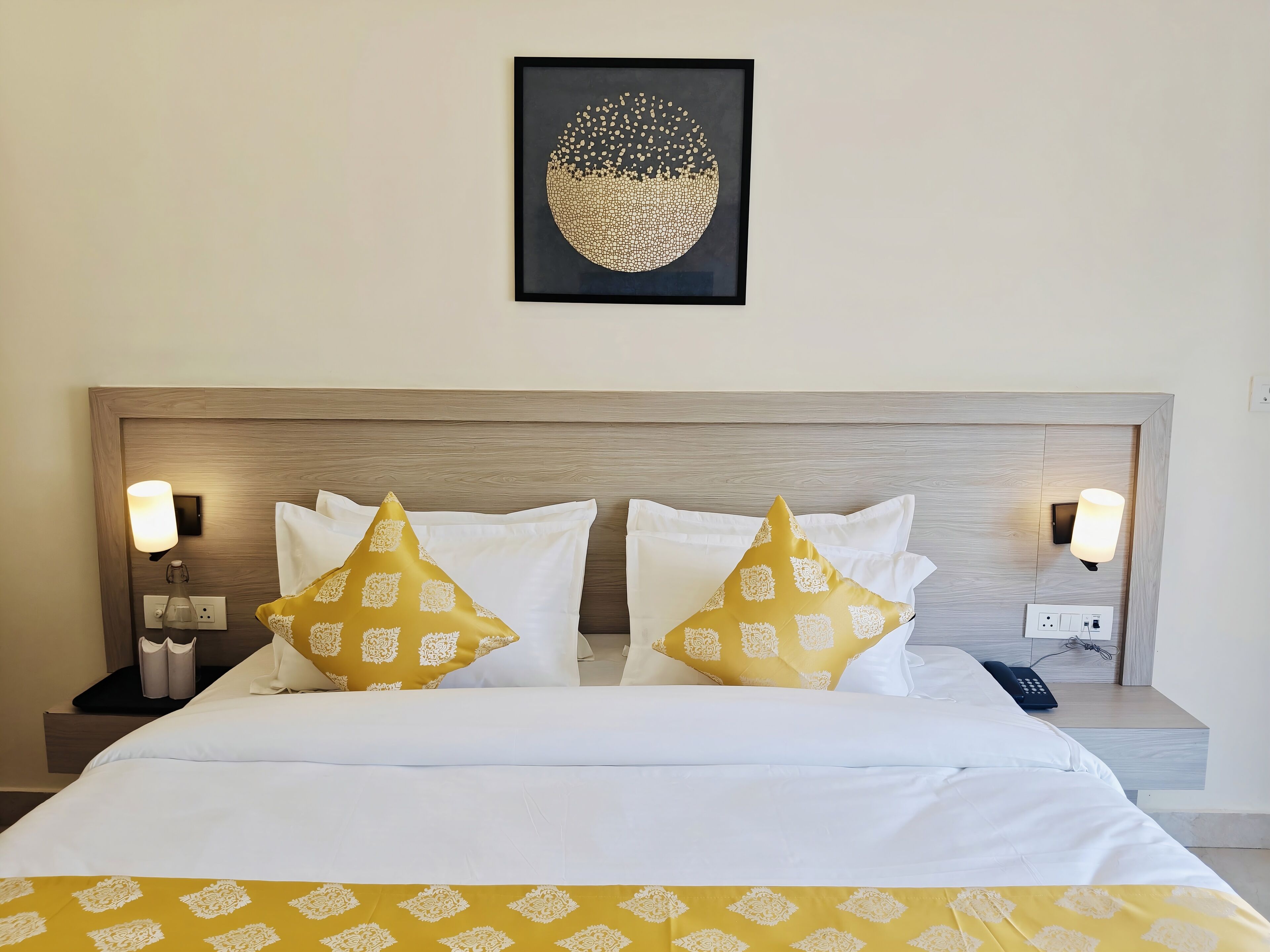 Ein Hotelzimmer mit einem Bett, zwei Nachttischen, einer Wandleuchte, einem gerahmten Kunstwerk und einer gemusterten Bettdecke.