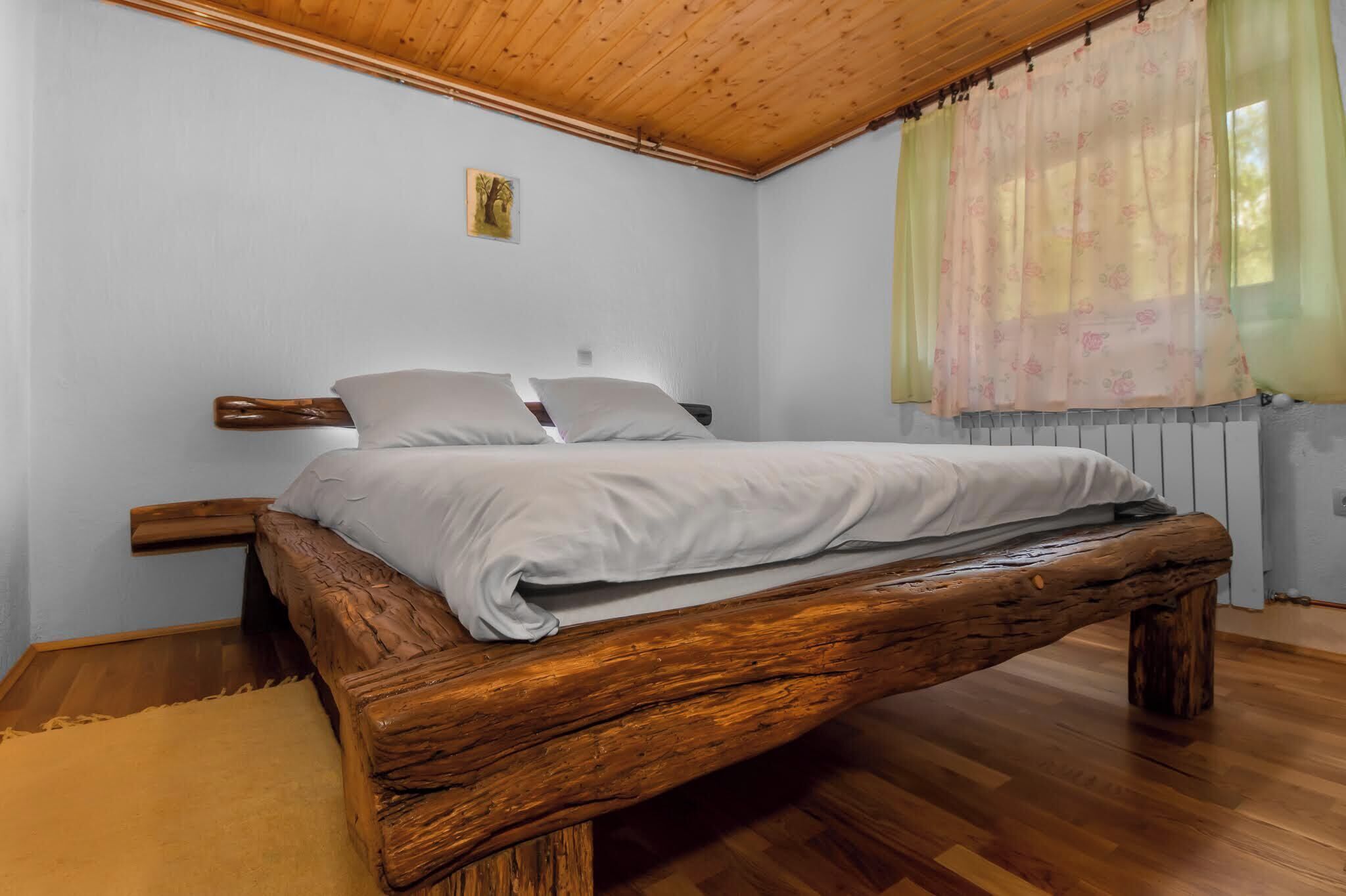 Un dormitorio con estructura de cama de madera, ropa de cama blanca, una mesita de noche pequeña y una ventana con cortinas translúcidas.