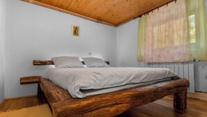 Un dormitorio con estructura de cama de madera, ropa de cama blanca, una mesita de noche pequeña y una ventana con cortinas translúcidas.
