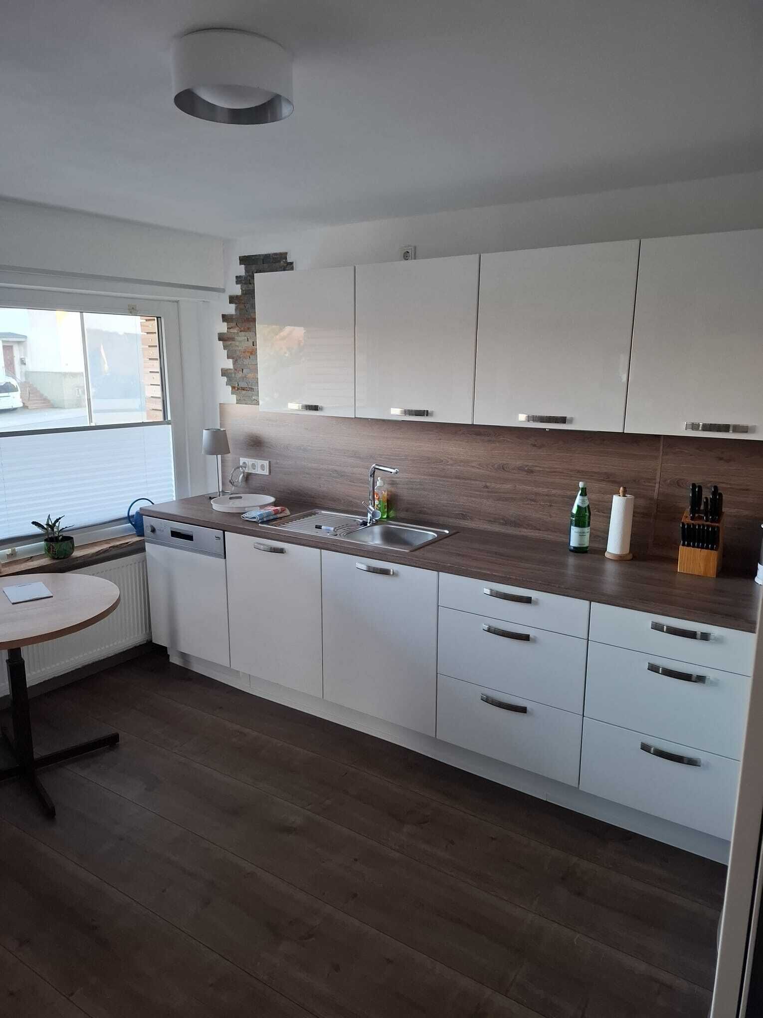 Une cuisine moderne avec des meubles blancs, un plan de travail en bois, un lave-vaisselle encastré et une fenêtre avec des stores.