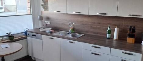 Une cuisine moderne avec des meubles blancs, un plan de travail en bois, un lave-vaisselle encastré et une fenêtre avec des stores.