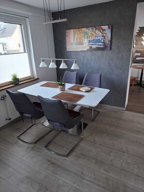 Ein Essbereich mit einem weißen Tisch, vier Stühlen, einer Topfpflanze und einem Bild an der Wand.