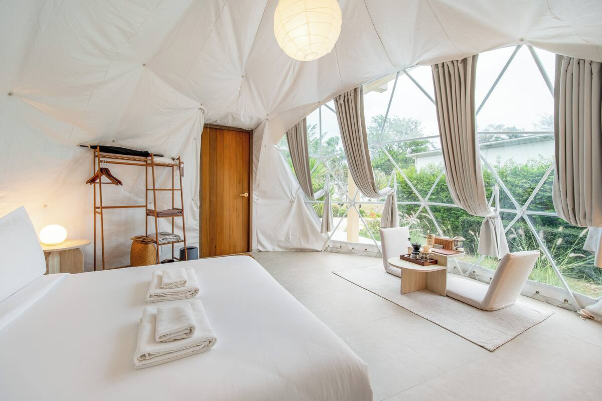 A tent-style room with a bed, a chair, a small table, and a view of greenery outside.