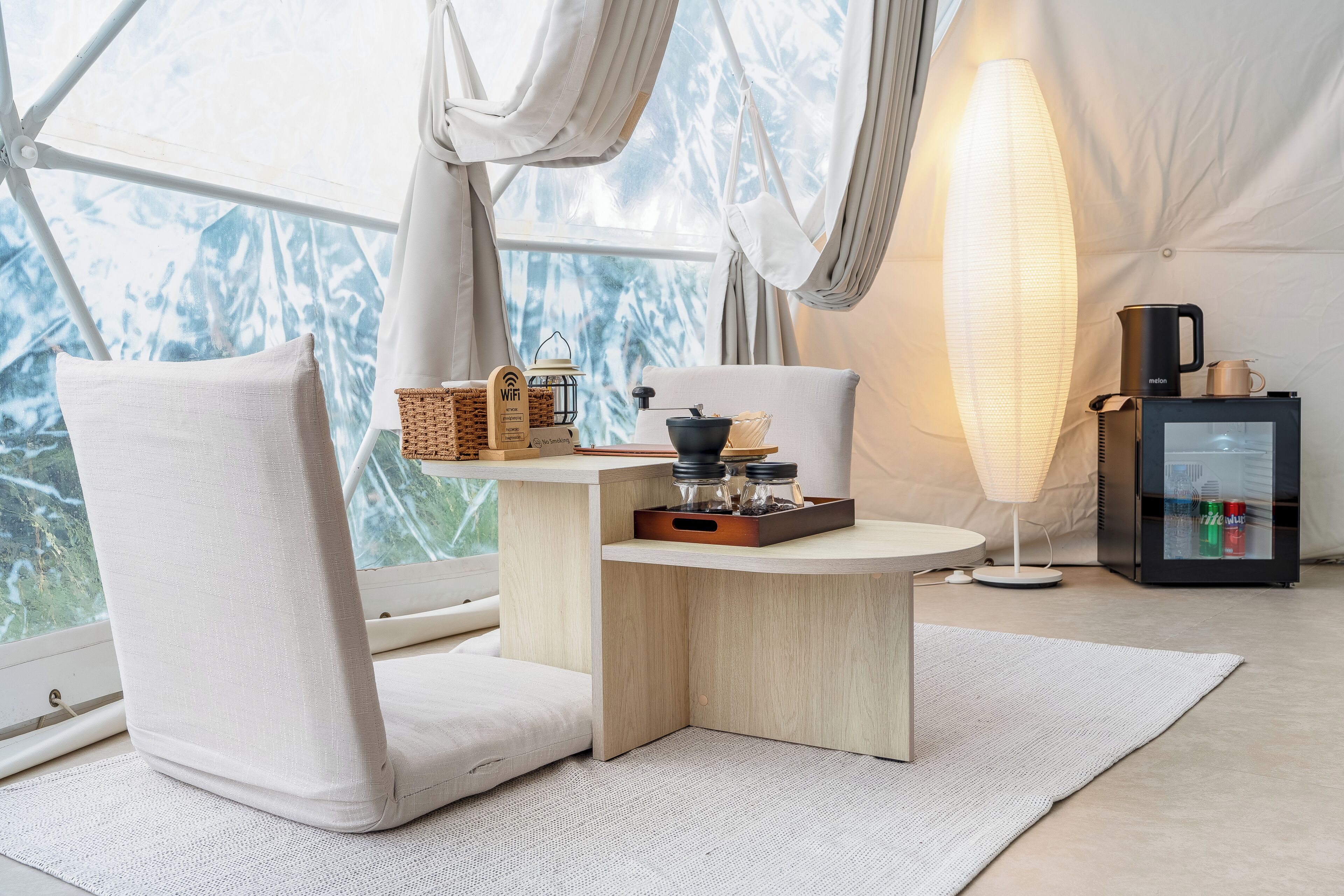 Une pièce confortable avec une grande fenêtre, une table avec une lampe, une chaise et un réfrigérateur.