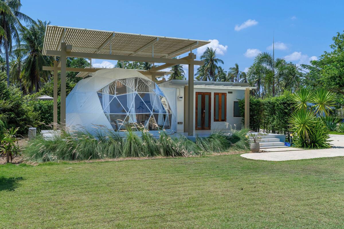A modern house with a large spherical glass structure, a wooden pergola, and tropical vegetation.