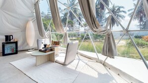 A tented room with a view of palm trees, a table with a lamp, and a chair. - Grood Glamping (Bang Saphan)