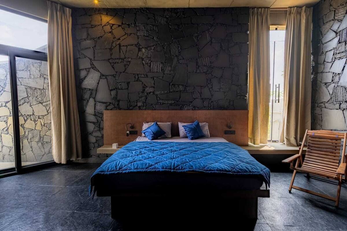 A bedroom with a stone wall, a bed with a blue quilt, a wooden headboard, and a window with curtains.