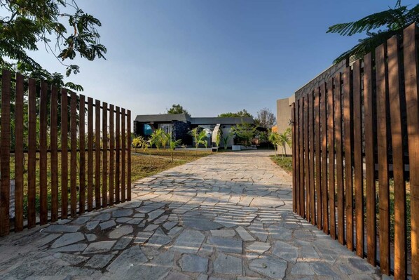 A stone pathway leading to a gated entrance with a modern building in the background. - Meraki (Shoolagiri)
