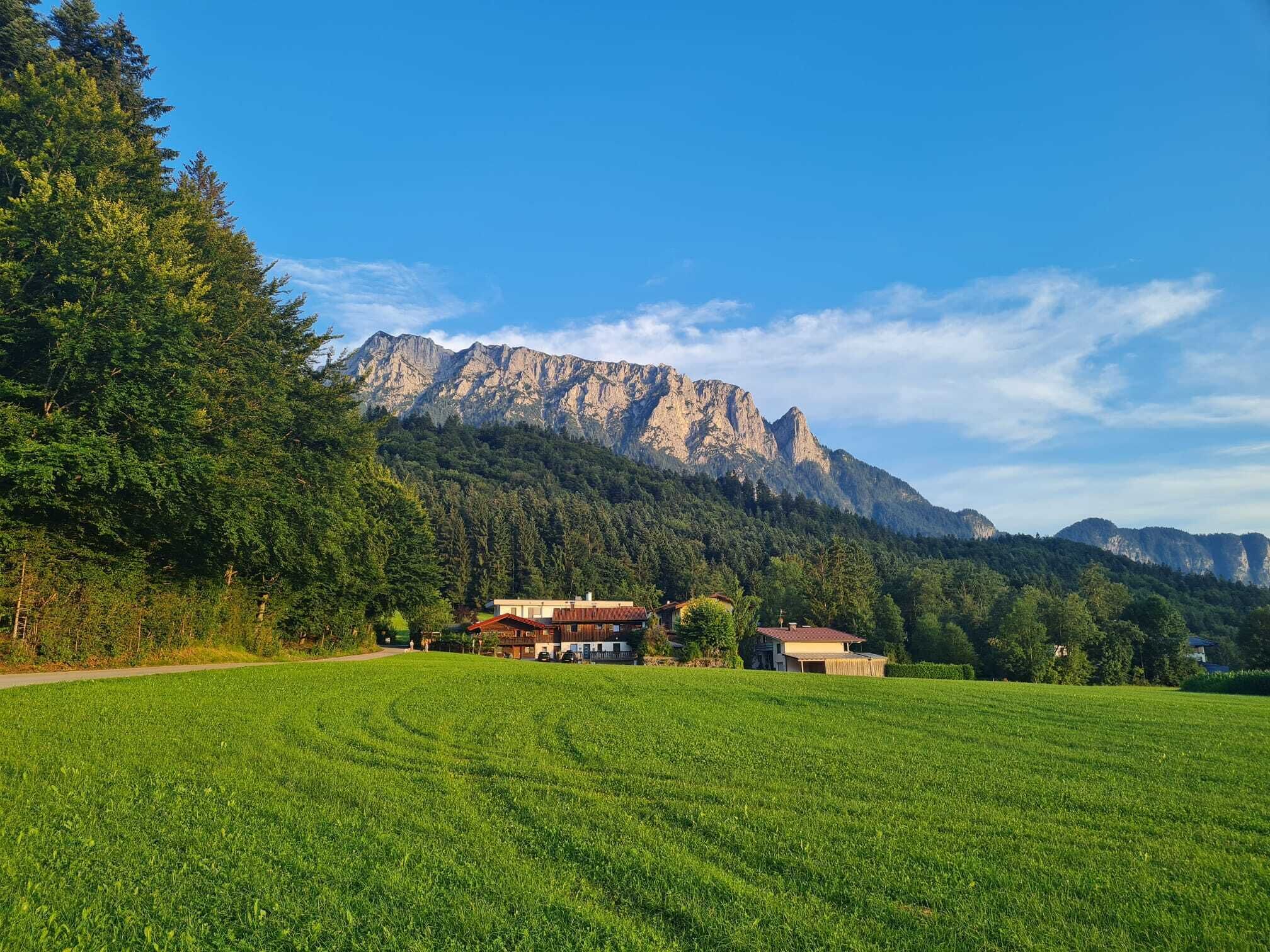 Un paysage rural avec une chaîne de montagnes en arrière-plan, un champ verdoyant et deux bâtiments, le tout sous un ciel dégagé.