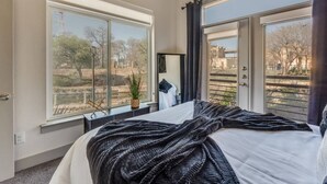 Une chambre avec une grande fenêtre, un lit, une table de chevet et une vue sur les arbres et les bâtiments.