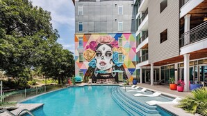 Une piscine agrémentée d’une fresque sur le bâtiment adjacent, entourée de verdure et bordée d’une architecture moderne.