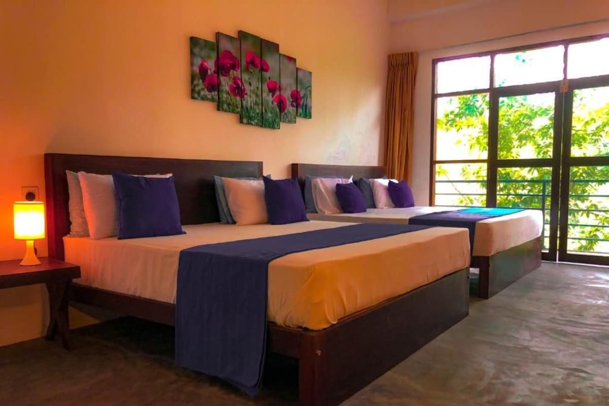 A hotel room with two beds, a nightstand with a lamp, a large window with a view of greenery, and a wall with a floral painting.