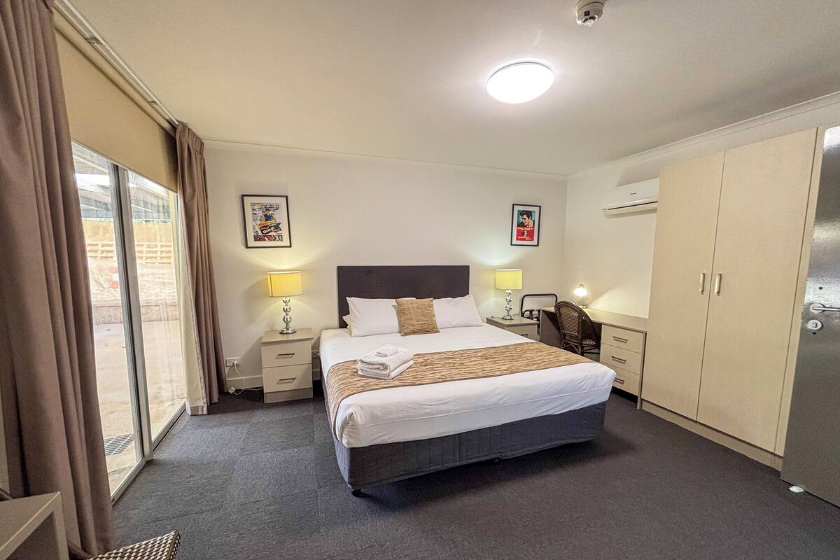 Une chambre d’hôtel comprenant un lit, deux tables de chevet avec des lampes, un bureau, une chaise, une armoire et une grande fenêtre avec des rideaux.