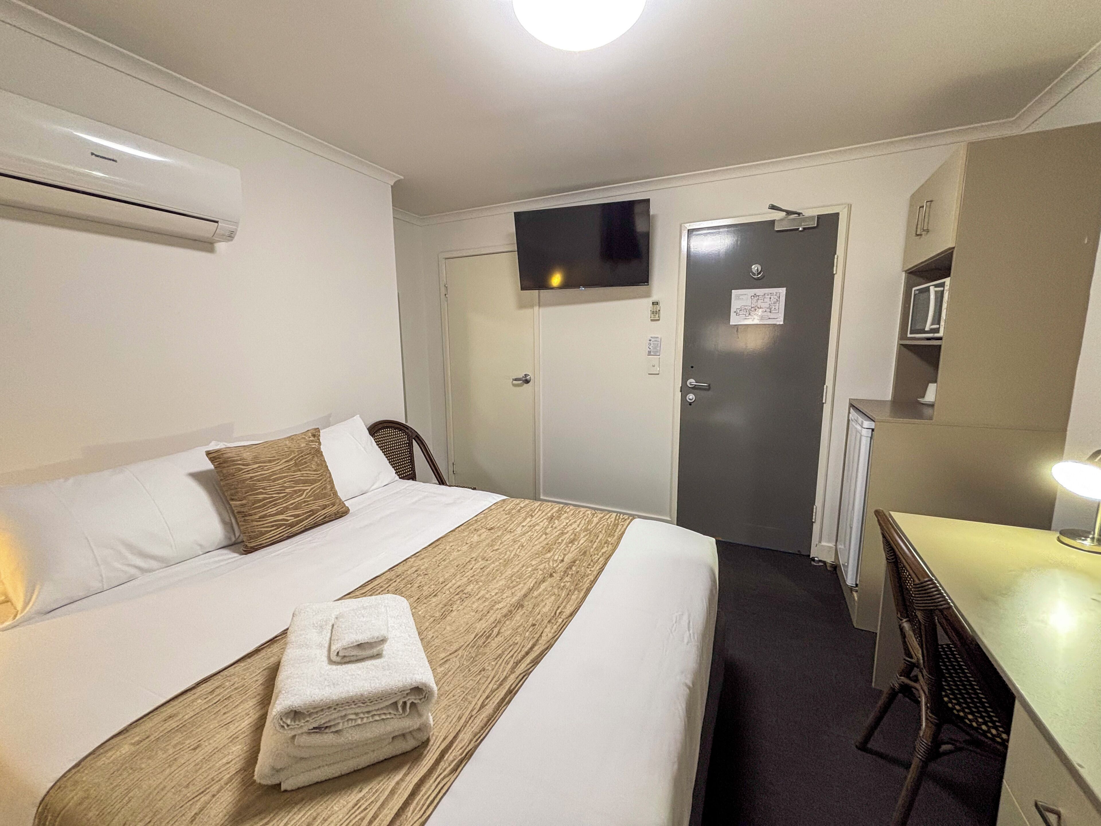 Une chambre d’hôtel équipée d’un lit, d’un bureau, d’une chaise, d’un climatiseur et d’une porte au-dessus de laquelle est fixée une télévision.