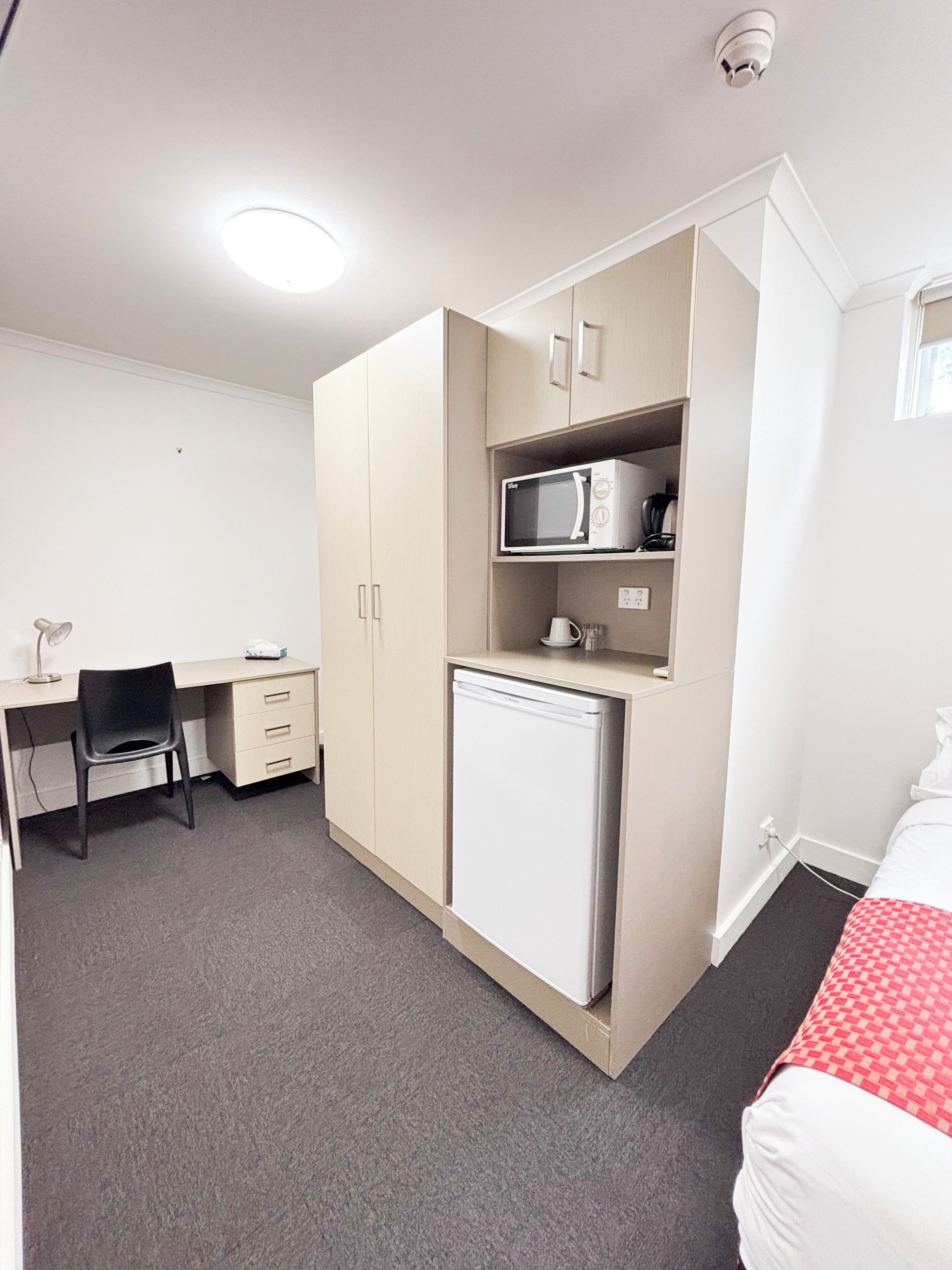 Habitación compacta con kitchenette, escritorio con silla y cama con manta de cuadros rojos.