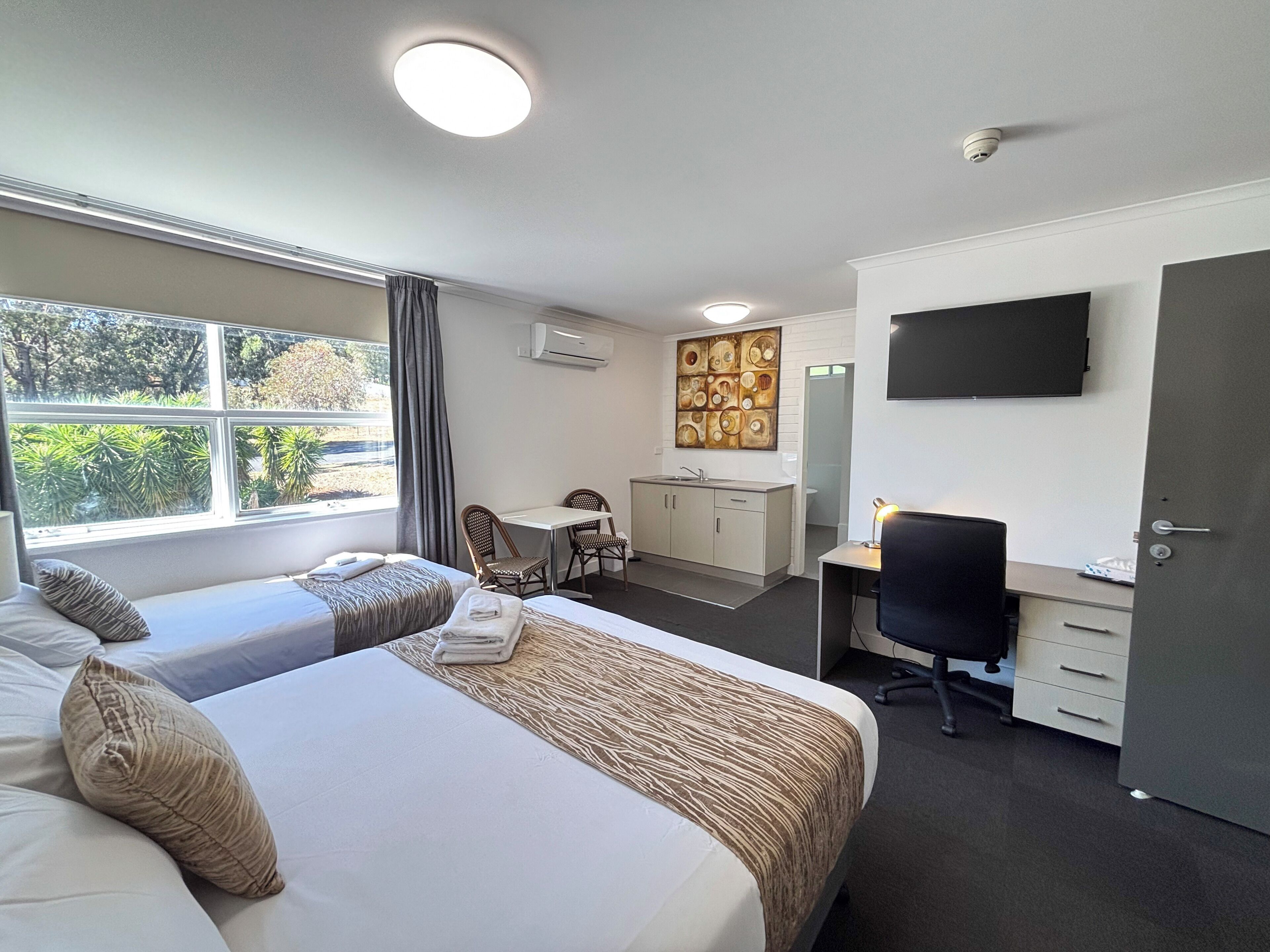 Quarto de hotel com duas camas, uma escrivaninha, uma cadeira, televisão de tela plana e janela com vista para árvores.