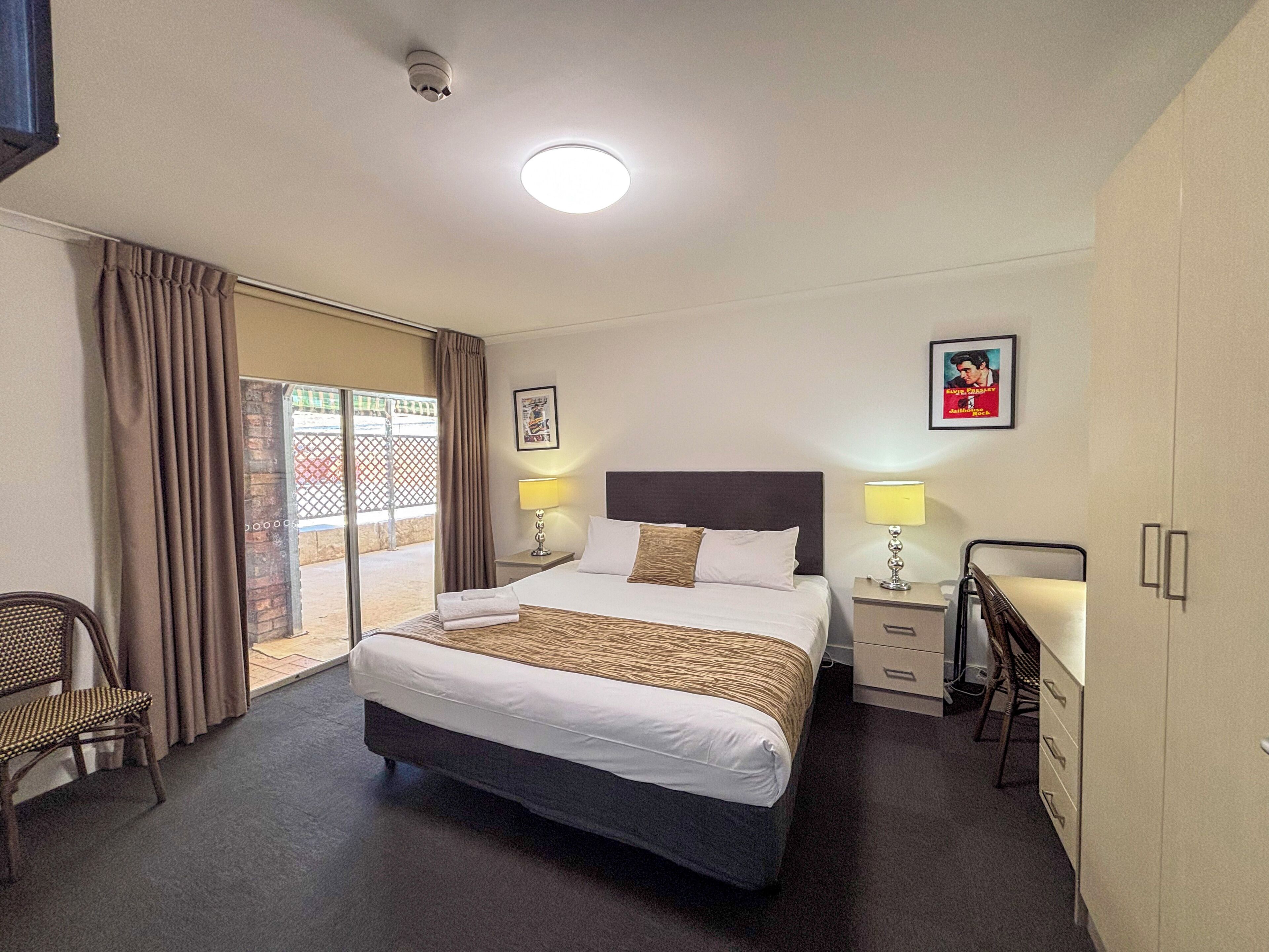 A hotel room with a bed, two bedside tables with lamps, a chair, a desk, and a sliding glass door to the outside.