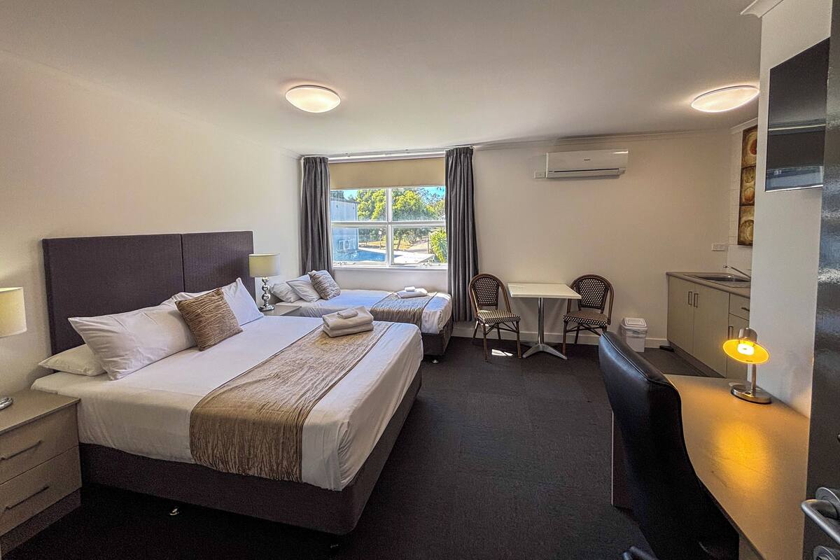 Une chambre d’hôtel avec deux lits, une petite table avec des chaises, un bureau avec une chaise, une fenêtre avec des rideaux et un appareil de climatisation.