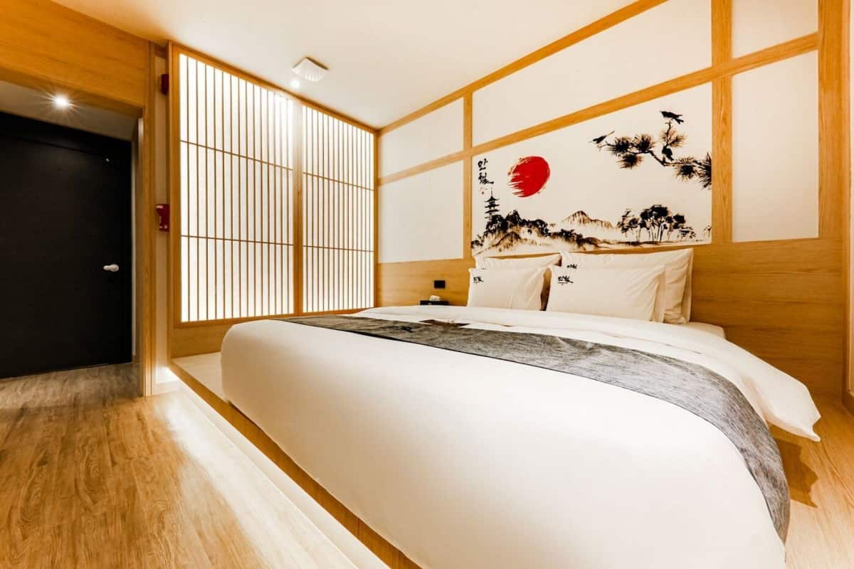A hotel room with a large bed, wooden headboard, shoji screens, and a traditional Japanese painting.