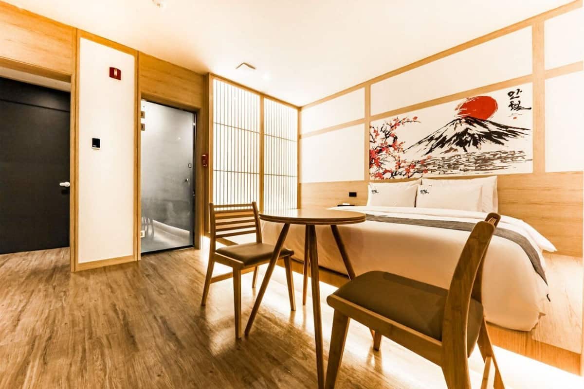 A hotel room with a wooden floor, a bed with white bedding, a small round table, two chairs, a sliding door, and a wall with a painting.
