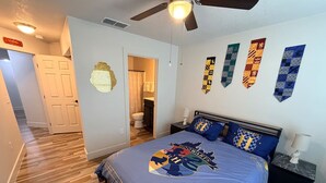 Quarto com cama, ventilador de teto, mesa de cabeceira e banheiro visível por uma porta aberta.