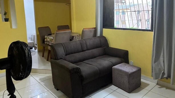 A living room with a sofa, ottoman, fan, dining table, chairs, and a window with curtains.