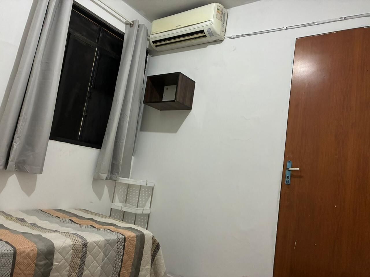 Ein Schlafzimmer mit einem Bett, einem Fenster mit Vorhängen, einer Klimaanlage, einer Holztür und einem kleinen Regal.