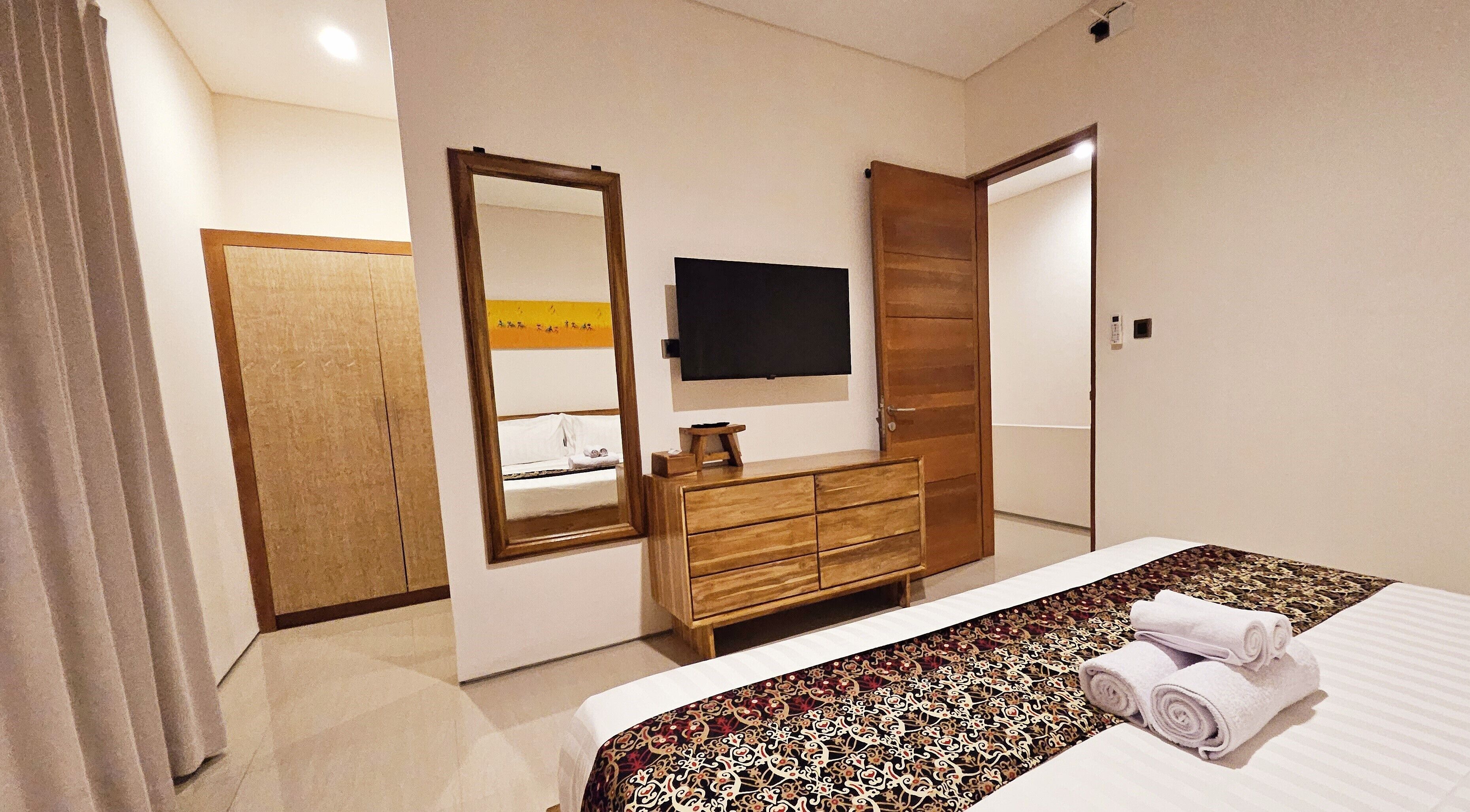 A hotel room with a bed, a wooden dresser, a mirror, a flat-screen TV, and a door.