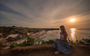 Une femme est assise sur un rocher, d’où elle a une vue sur une plage. On aperçoit un bateau et un bâtiment au loin.