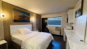 Una habitación de hotel con una cama grande, un escritorio con silla, una ventana con persianas, un cuadro en la pared y una kitchenette pequeña.