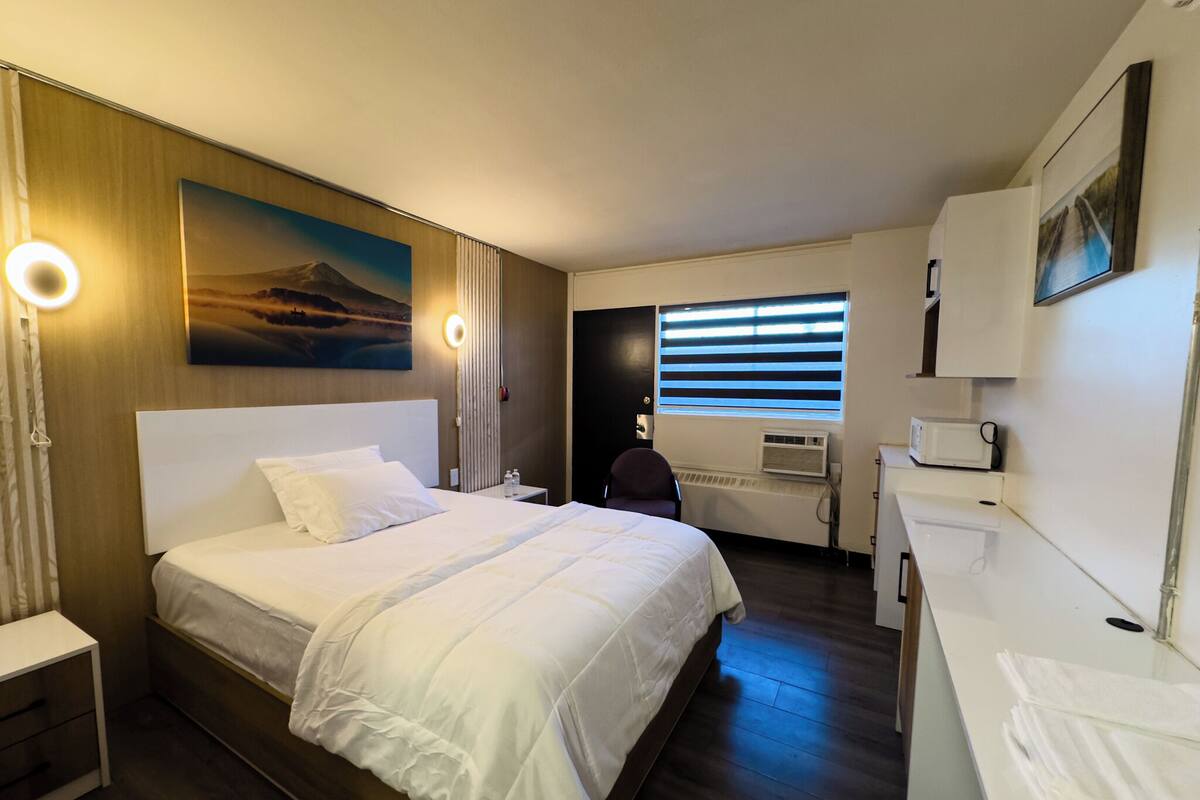 A hotel room with a large bed, a desk with a chair, a window with blinds, a painting on the wall, and a small kitchenette.