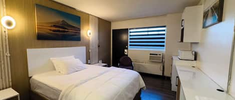 A hotel room with a large bed, a desk with a chair, a window with blinds, a painting on the wall, and a small kitchenette.