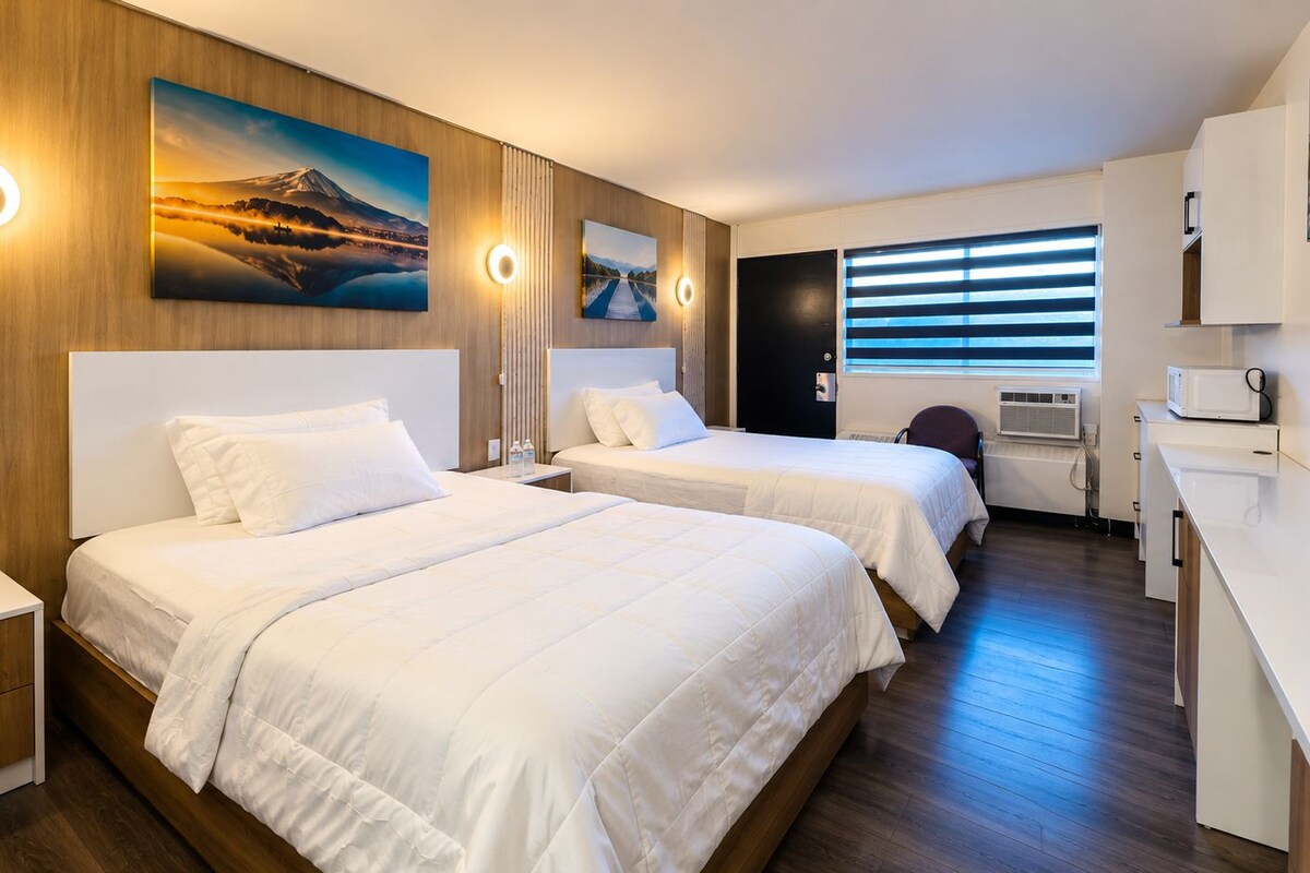 A hotel room with two beds, a desk, and a chair. There are wall-mounted lights and artwork.