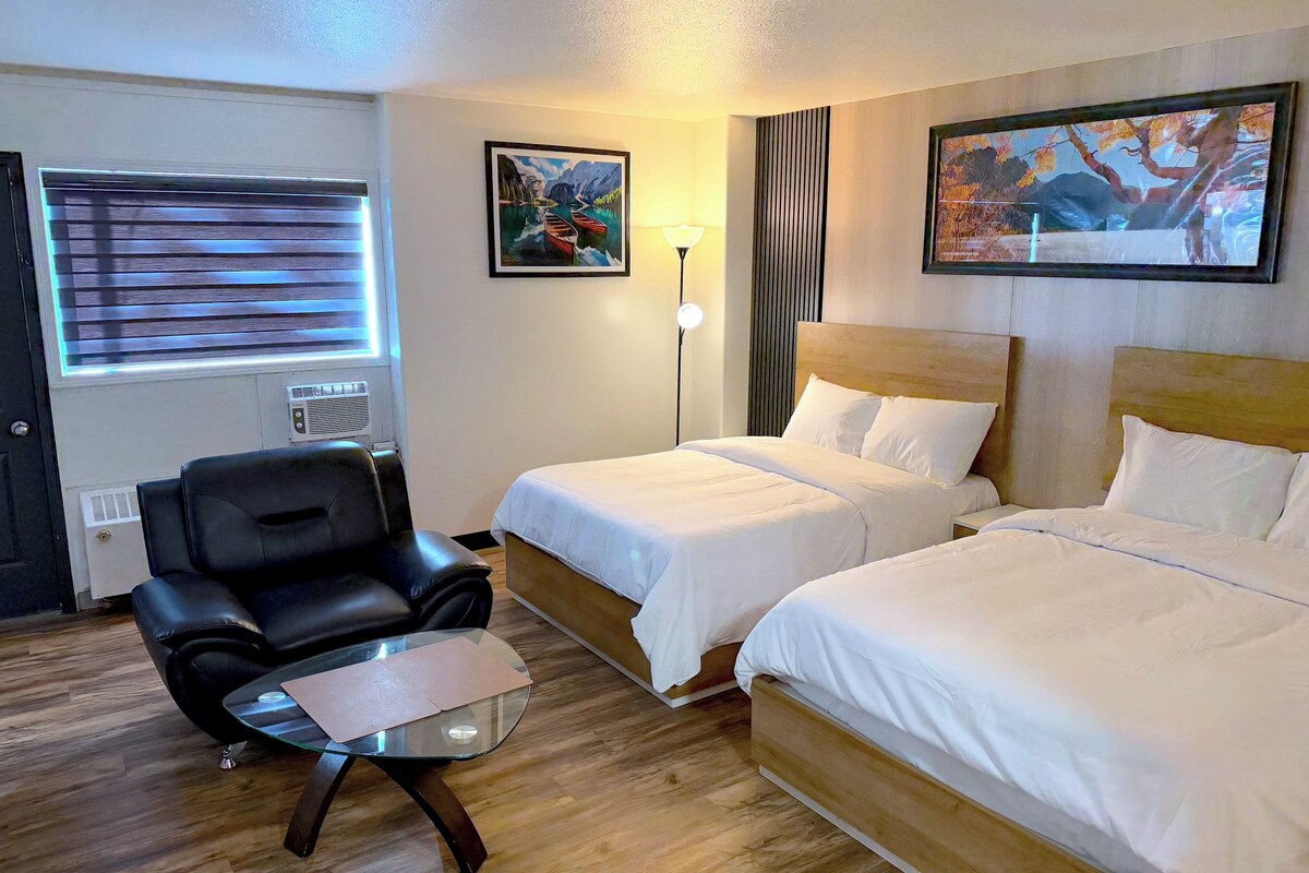 Quarto de hotel com duas camas, uma escrivaninha com cadeira, janela com persianas e dois quadros na parede.