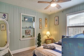 Une chambre à coucher comprenant un lit, une table de chevet avec une lampe, une fenêtre avec des stores, une étagère fixée au mur avec des objets décoratifs et un ventilateur de plafond.