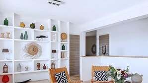 Una stanza con un tavolo di legno, due sedie in vimini con cuscini decorati, un ripiano bianco con diversi oggetti ornamentali e un grande specchio a parete.