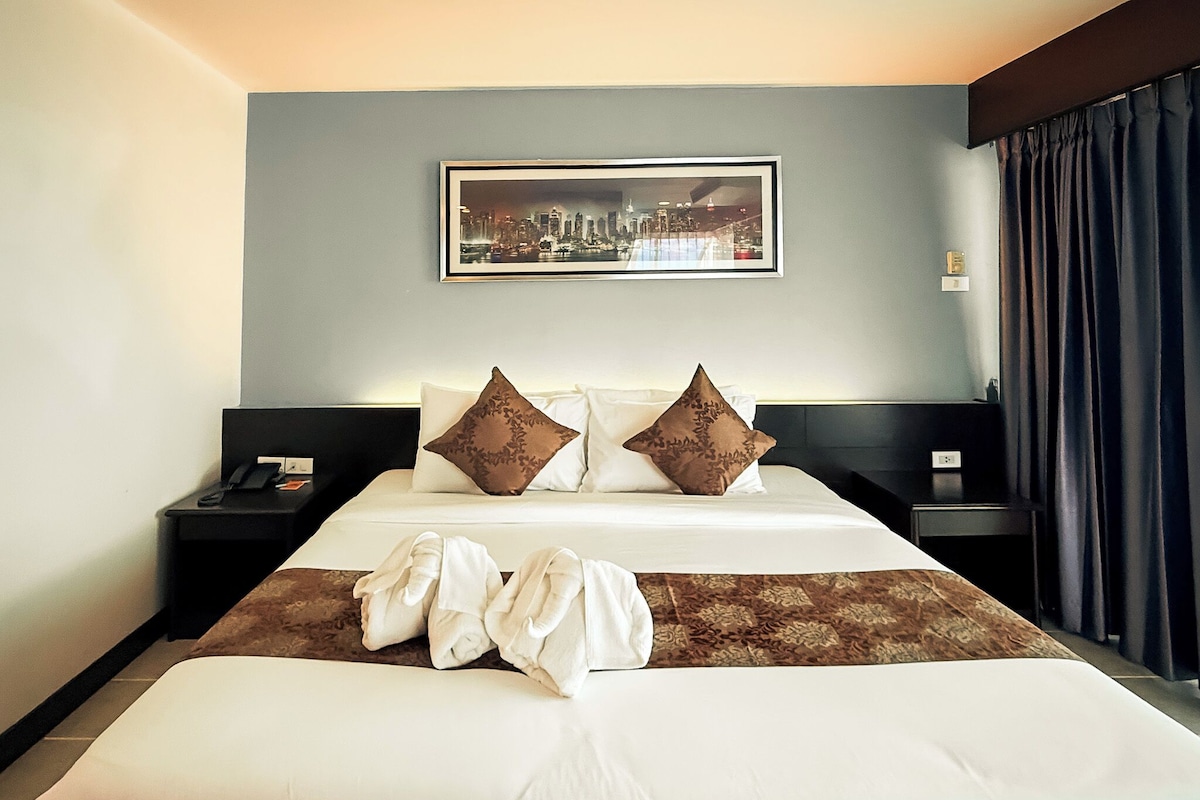 A hotel room with a large bed, two nightstands, a framed cityscape picture on the wall, and a dark curtain on the window.