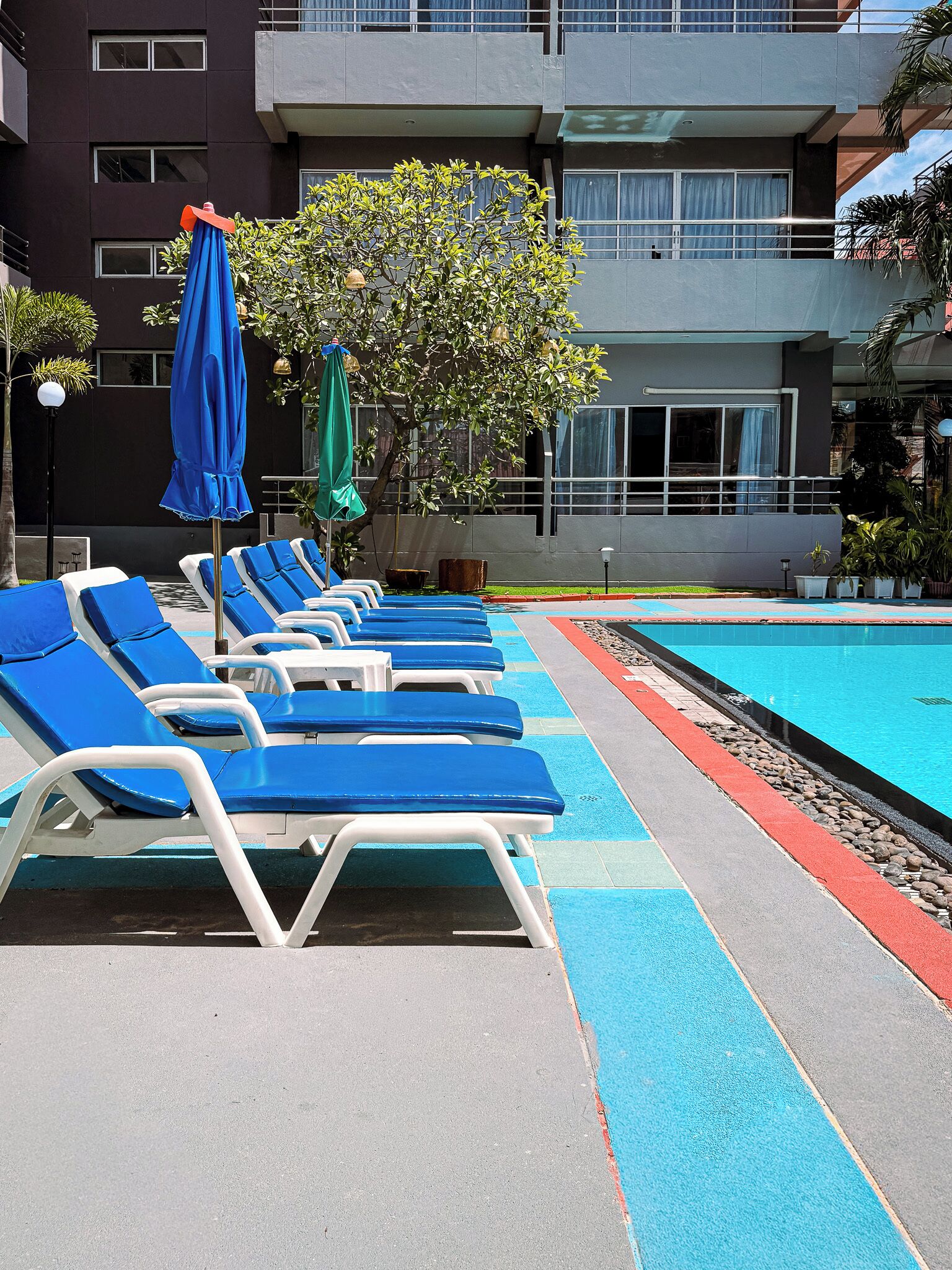A poolside area with lounge chairs and umbrellas, a swimming pool, and a building with multiple windows and balconies.