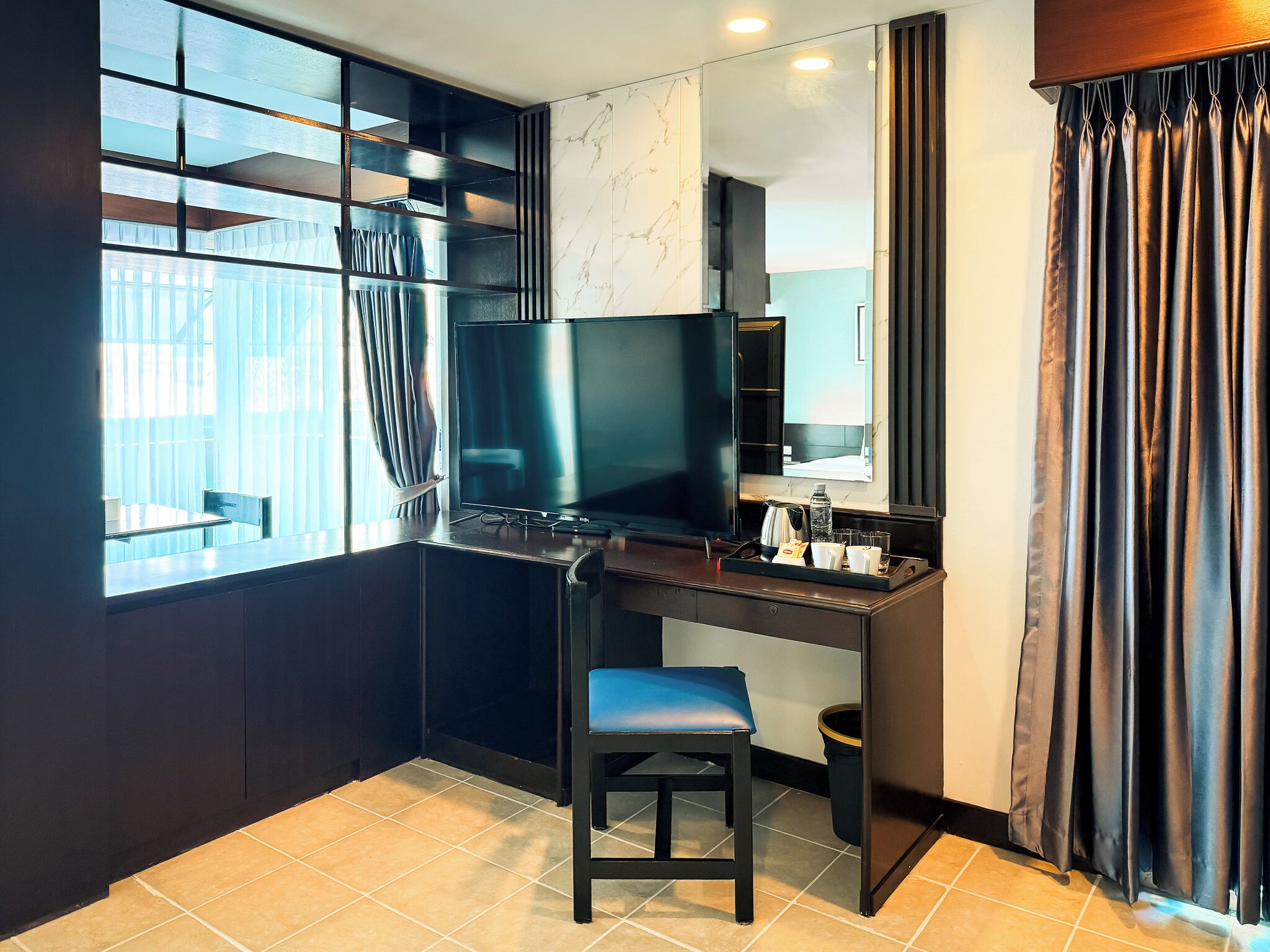 A hotel room with a desk, a television, a chair, a mirror, and a window with curtains.